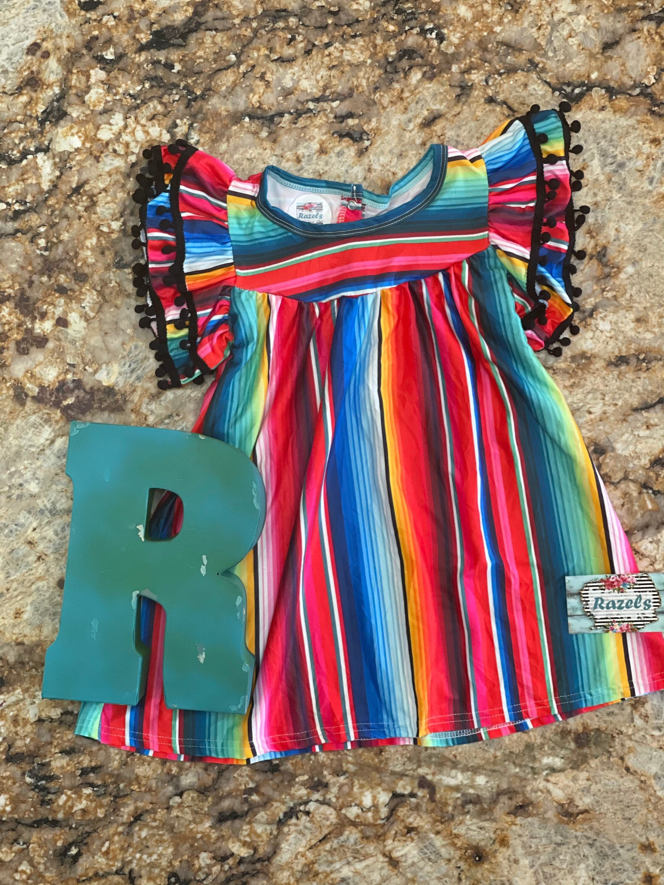 The girls&#39; fiesta party dress features a bright serape stripe throughout and pom-pom cap sleeves.