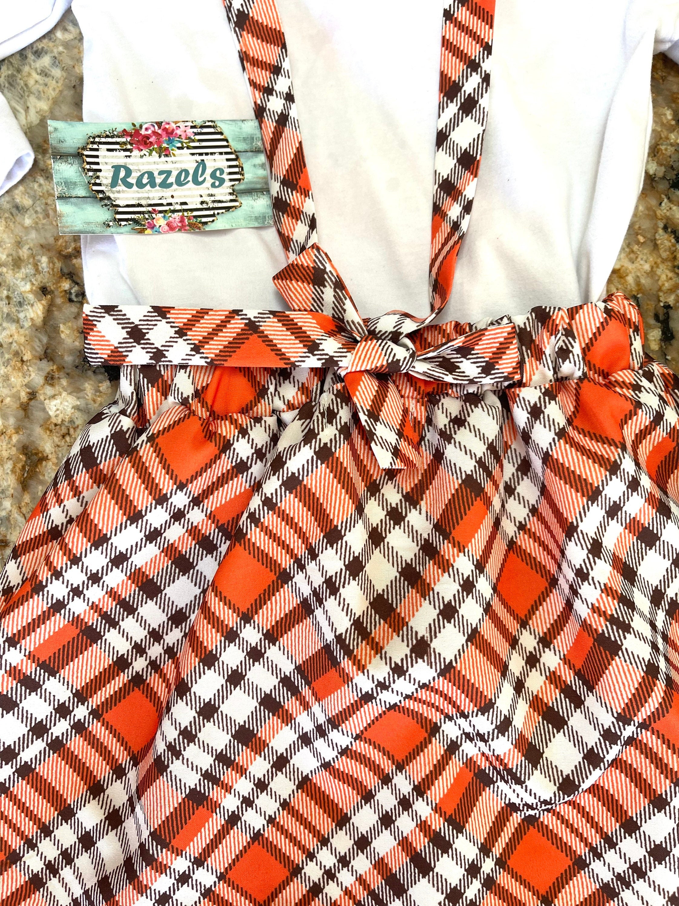 Girls Fall Thankful Outfit. The Fall Plaid Suspender Skirt features an orange, brown, and white plaid pattern with suspenders. The shirt boasts ruffled shoulders, long sleeves, and a glittering gold pumpkin with the word thankful in the center.