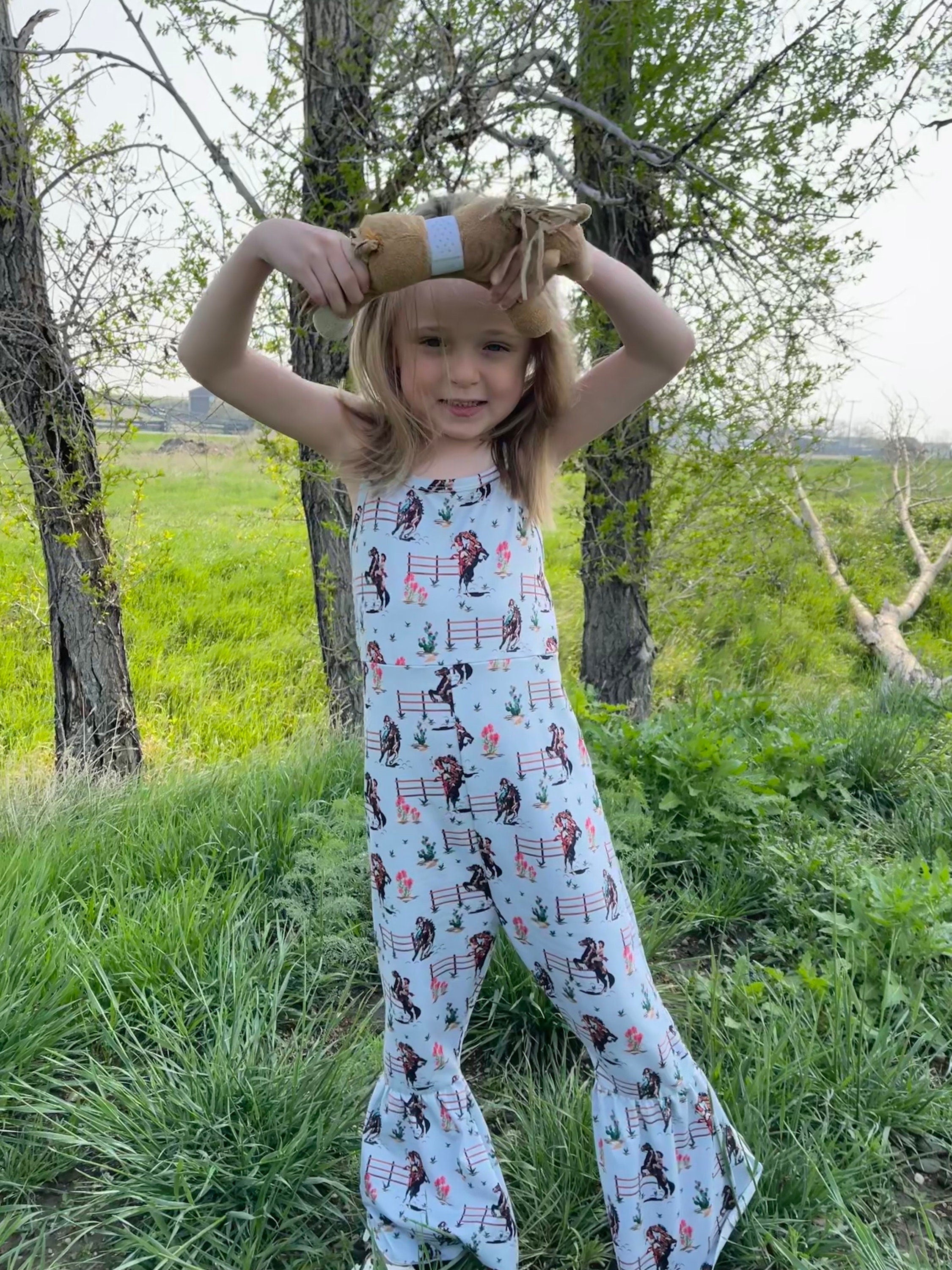 Jernie modeling Western Cowgirl Print Jumpsuit in Blue. This fun jumpsuit features a vintage cowgirl print of bucking and rearing horses and their best cowgirls.  Legs have a flare/bell bottom hem and spaghetti strap. An easy-peasy outfit!