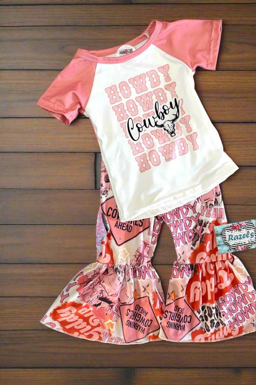 girls HowdyCowboy Bell Bottom Outfit. T-shirt is baseball style with short pink sleeves and Howdy Cowboy scrolled across the front. Pants are pink bell bottom with Howdy, Warning Cowgirl ahead, stars,horses, cowprint western print