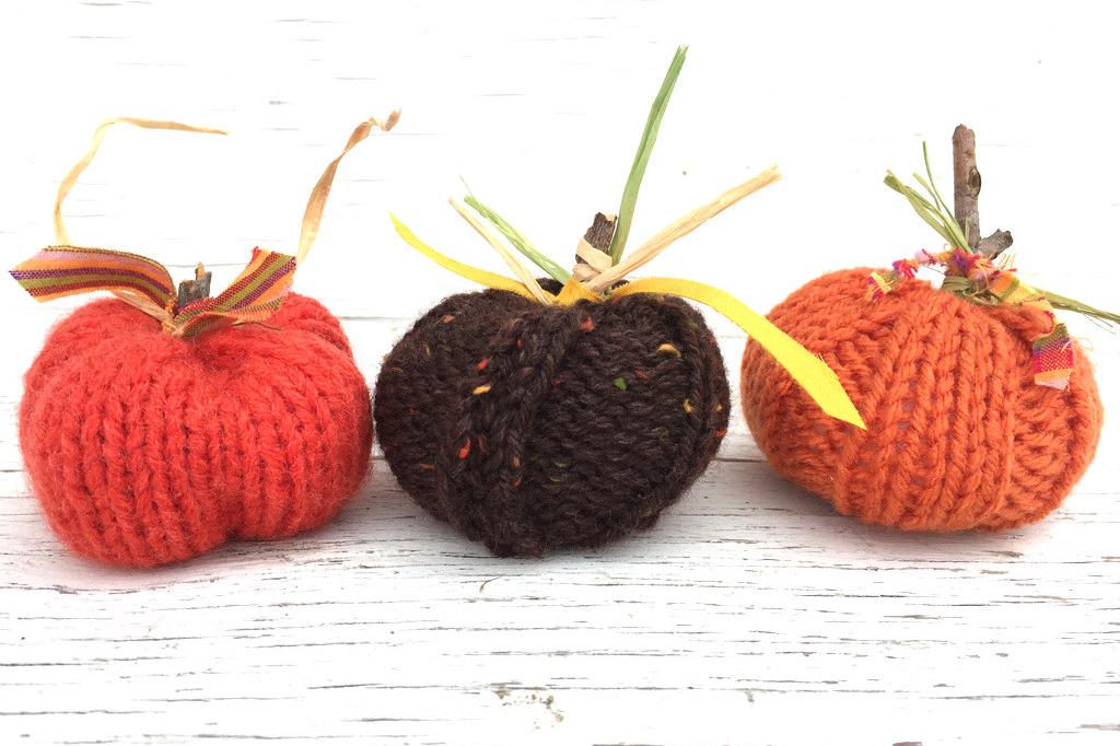 picture of knit pumpkins varying in size from small to large. Pumpkins are hand-knit with a ribbed appearance. The larger pumpkins have knit vines and leaves, while the smaller feature sticks with correlating fabric leaves.