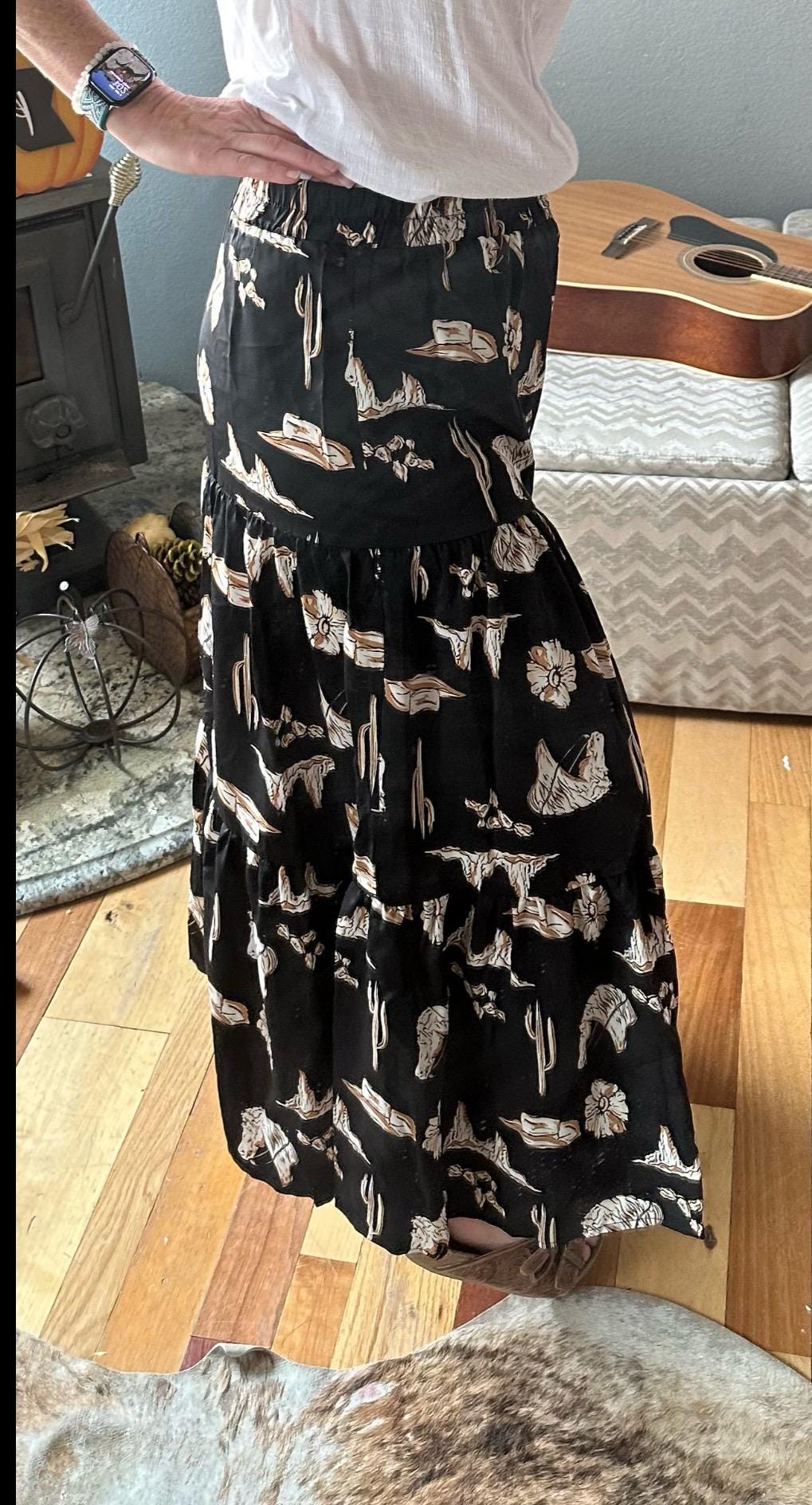 The Fall Women&#39;s Western BOHO maxi skirt features a charming western print with horses, cacti, flowers, and cowboy hats. It has a high-waisted design and a flowing silhouette in a fall color combo of black, brown, tan, and cream.