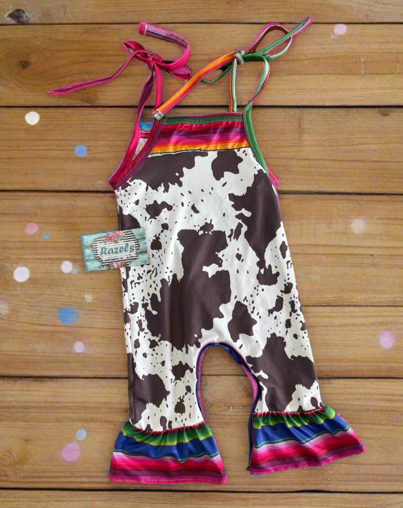 Cowprint Serape Jumpsuit—This stylish Cowprint Outfit features a bold cowprint design complemented by fun pink, blue, and green serape stripes on the neckline, shoulder tie straps, and ruffle hem. Easy 1 piece Cowgirl outfit. Ships FREE with $35+
