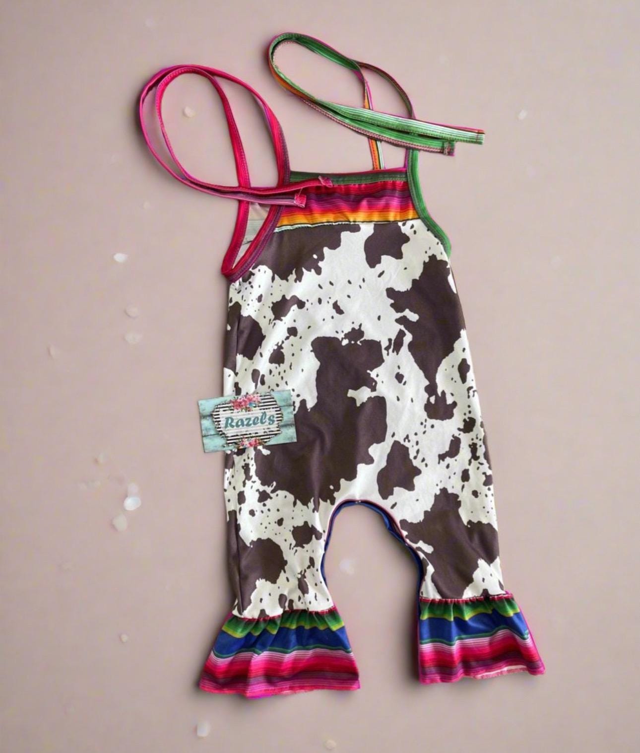 Cowprint Serape Jumpsuit—This stylish Cowprint Outfit features a bold cowprint design complemented by fun pink, blue, and green serape stripes on the neckline, shoulder tie straps, and ruffle hem. Easy 1 piece Cowgirl outfit. Ships FREE with $35+