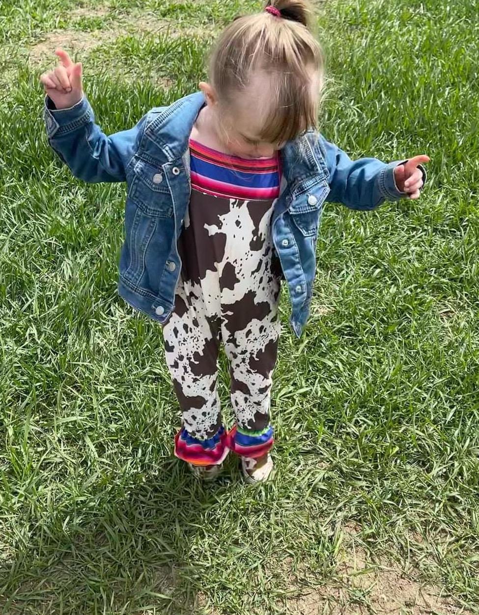 Cowprint Serape Jumpsuit—This stylish Cowprint Outfit features a bold cowprint design complemented by fun pink, blue, and green serape stripes on the neckline, shoulder tie straps, and ruffle hem. Easy 1 piece Cowgirl outfit. Ships FREE with $35+