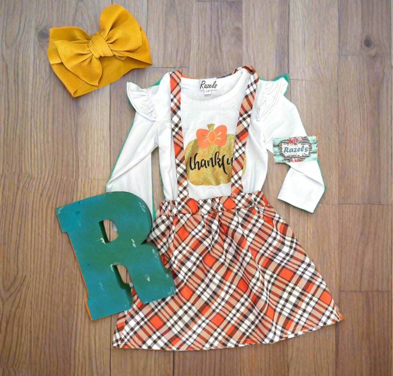 Girls Fall Thankful Outfit. The Fall Plaid Suspender Skirt features an orange, brown, and white plaid pattern with suspenders. The shirt boasts ruffled shoulders, long sleeves, and a glittering gold pumpkin with the word thankful in the center.