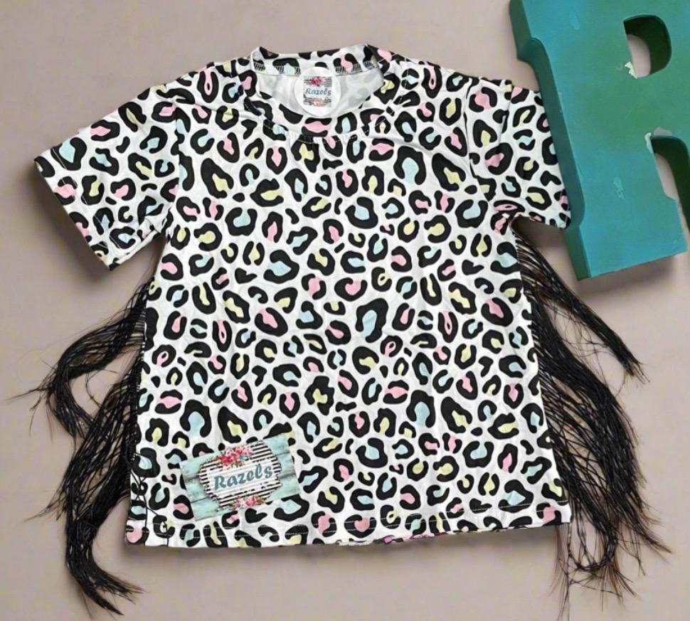 Girls Cheetah Print Fringe T-Shirt Dress: Multicolor cheetah print with pink, yellow, and blue highlighted by black. Adorned with black side fringe down both sides of the dress for a comfy, fun, and adorable look!