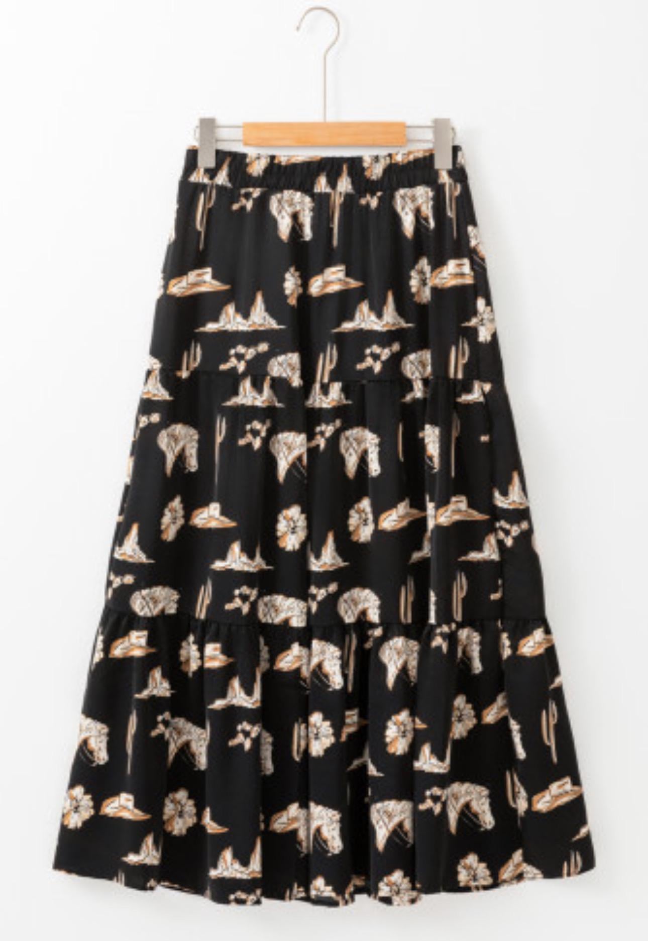 The Fall Women&#39;s Western BOHO maxi skirt features a charming western print with horses, cacti, flowers, and cowboy hats. It has a high-waisted design and a flowing silhouette in a fall color combo of black, brown, tan, and cream.