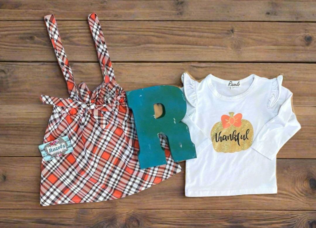 Girls Fall Thankful Outfit. The Fall Plaid Suspender Skirt features an orange, brown, and white plaid pattern with suspenders. The shirt boasts ruffled shoulders, long sleeves, and a glittering gold pumpkin with the word thankful in the center.