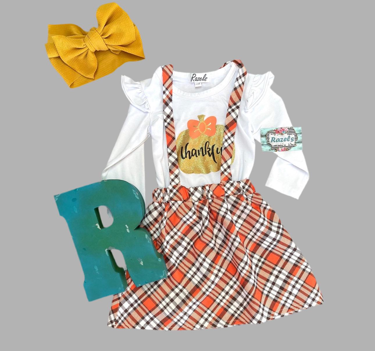 Girls Fall Thankful Outfit. The Fall Plaid Suspender Skirt features an orange, brown, and white plaid pattern with suspenders. The shirt boasts ruffled shoulders, long sleeves, and a glittering gold pumpkin with the word thankful in the center.