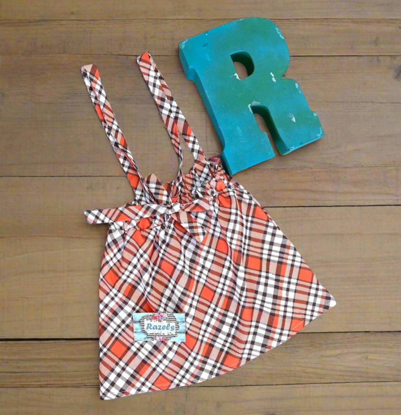 Girls Fall Thankful Outfit. The Fall Plaid Suspender Skirt features an orange, brown, and white plaid pattern with suspenders. The shirt boasts ruffled shoulders, long sleeves, and a glittering gold pumpkin with the word thankful in the center.