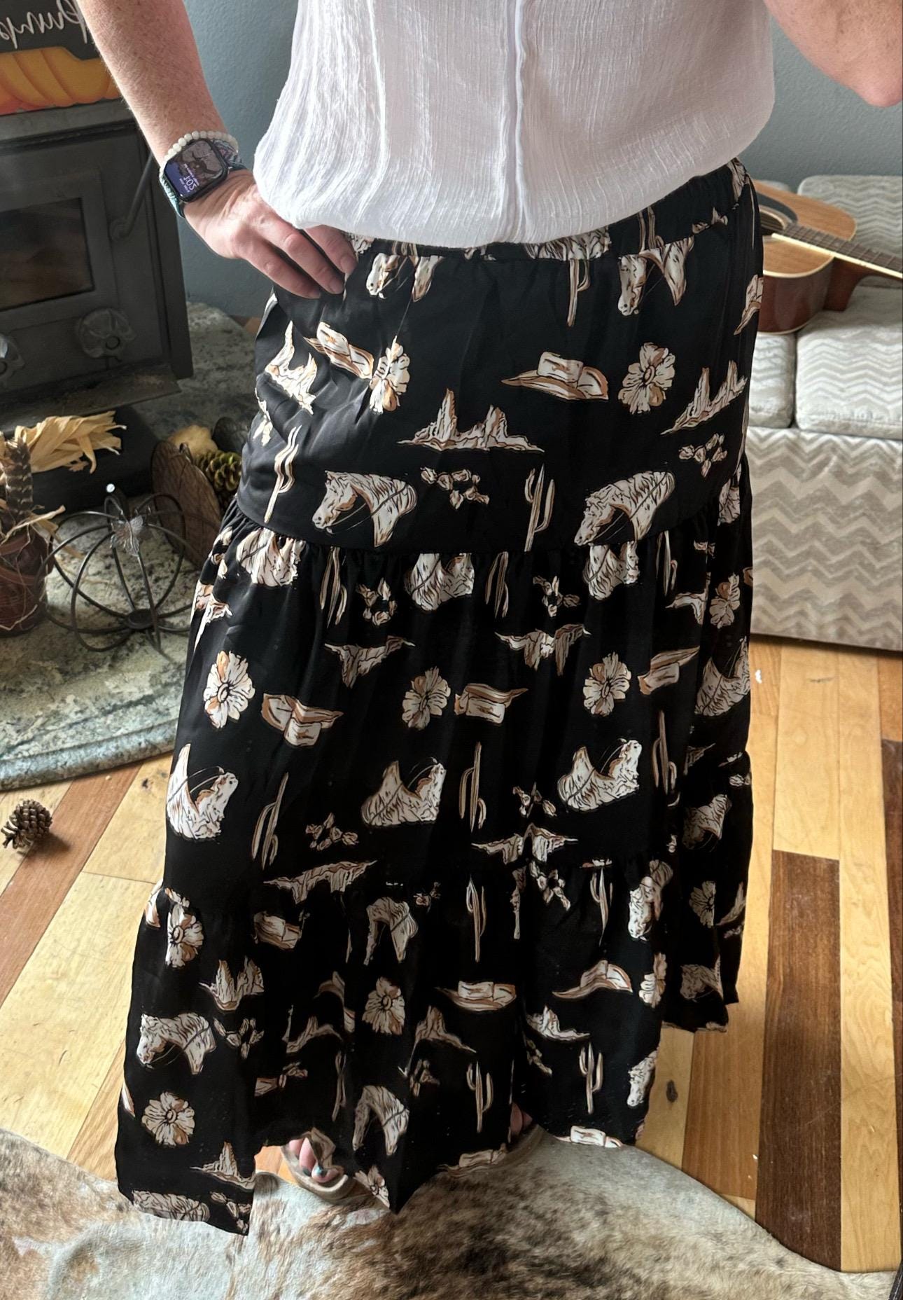 The Fall Women&#39;s Western BOHO maxi skirt features a charming western print with horses, cacti, flowers, and cowboy hats. It has a high-waisted design and a flowing silhouette in a fall color combo of black, brown, tan, and cream.