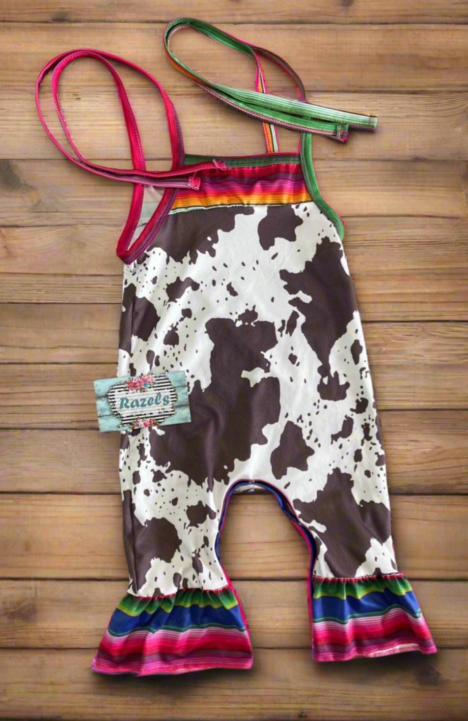 Cowprint Serape Jumpsuit—This stylish Cowprint Outfit features a bold cowprint design complemented by fun pink, blue, and green serape stripes on the neckline, shoulder tie straps, and ruffle hem. Easy 1 piece Cowgirl outfit. Ships FREE with $35+
