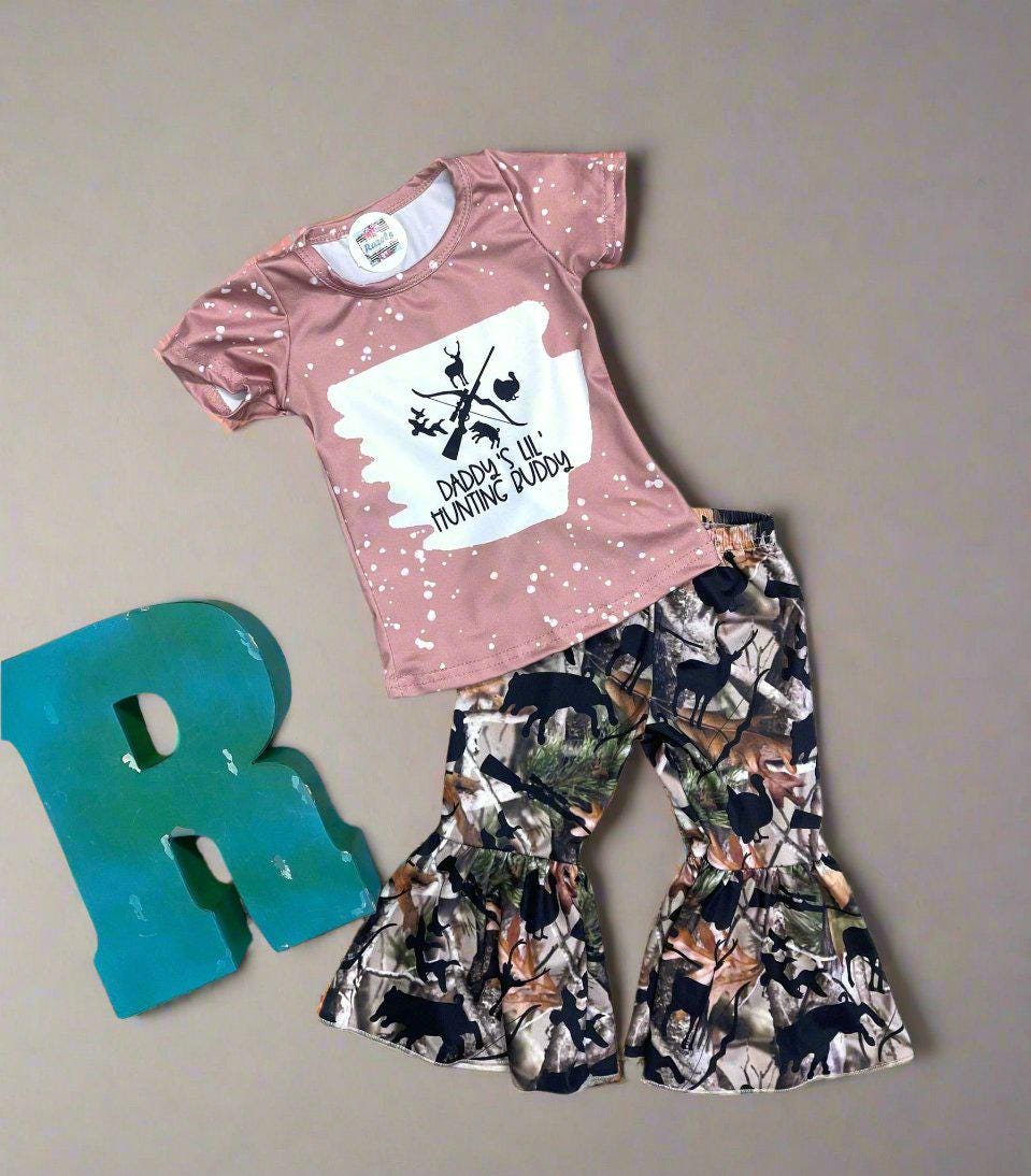 Girls&#39; &quot;Daddy&#39;s Lil Hunting Buddy&quot; bell-bottom outfit. The shirt is a pink t-shirt with a bleach-splattered style featuring a hunting motif of deer, turkey, and pig, as well as a bow and rifle, accompanied by camo print bell-bottom pants.