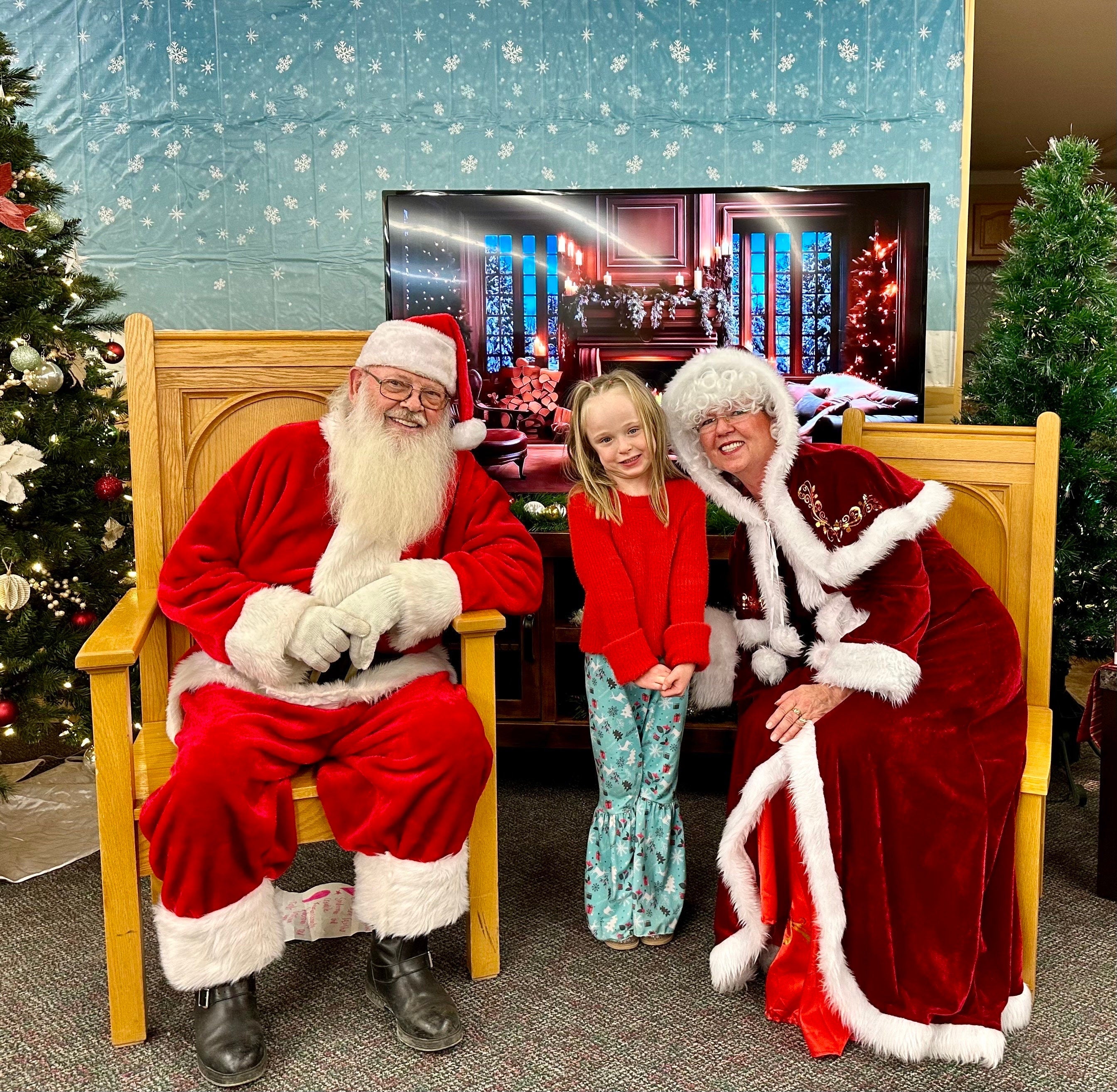 May include: Santa Claus wearing a red suit with white trim and a white beard is sitting in a wooden chair. A girl wearing a red sweater and blue pants is standing next to Santa. A woman wearing a red dress with white trim and a white hat is sitting in a wooden chair next to the girl.