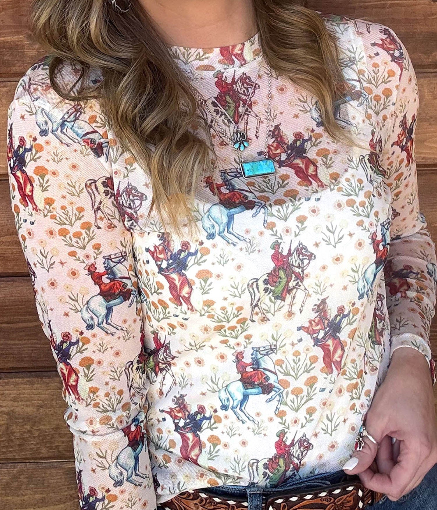 A woman wearing a long-sleeve white top with a colorful cowboy and rider print, paired with a brown western belt and blue jeans, standing against a rustic wooden wall.