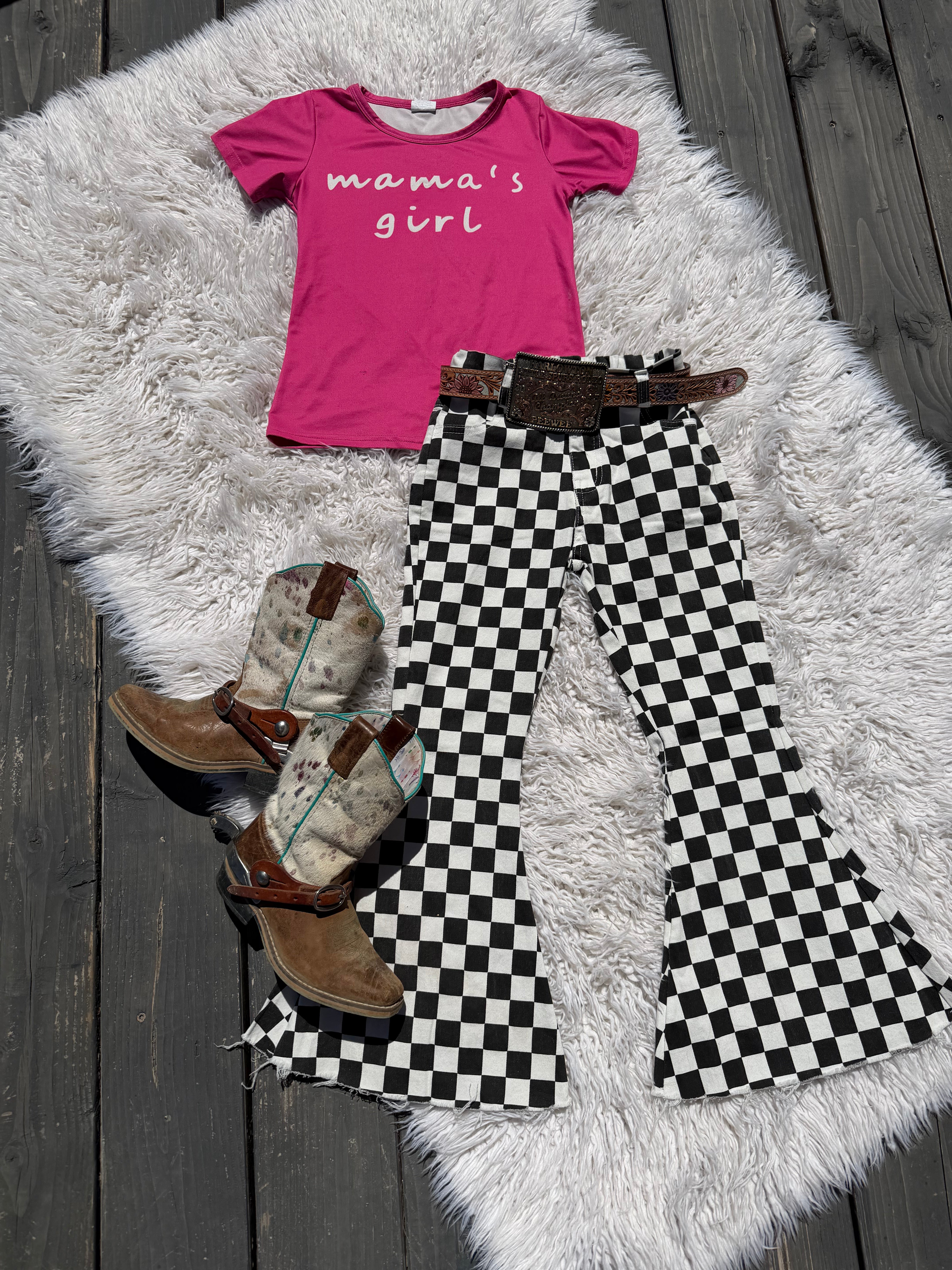 A styled flat-lay outfit for a child featuring a bright pink t-shirt with 'mama's girl' in white script, black and white checkered flared pants, a brown leather belt, and brown western boots, all arranged on a white faux fur rug over dark gray wooden planks.