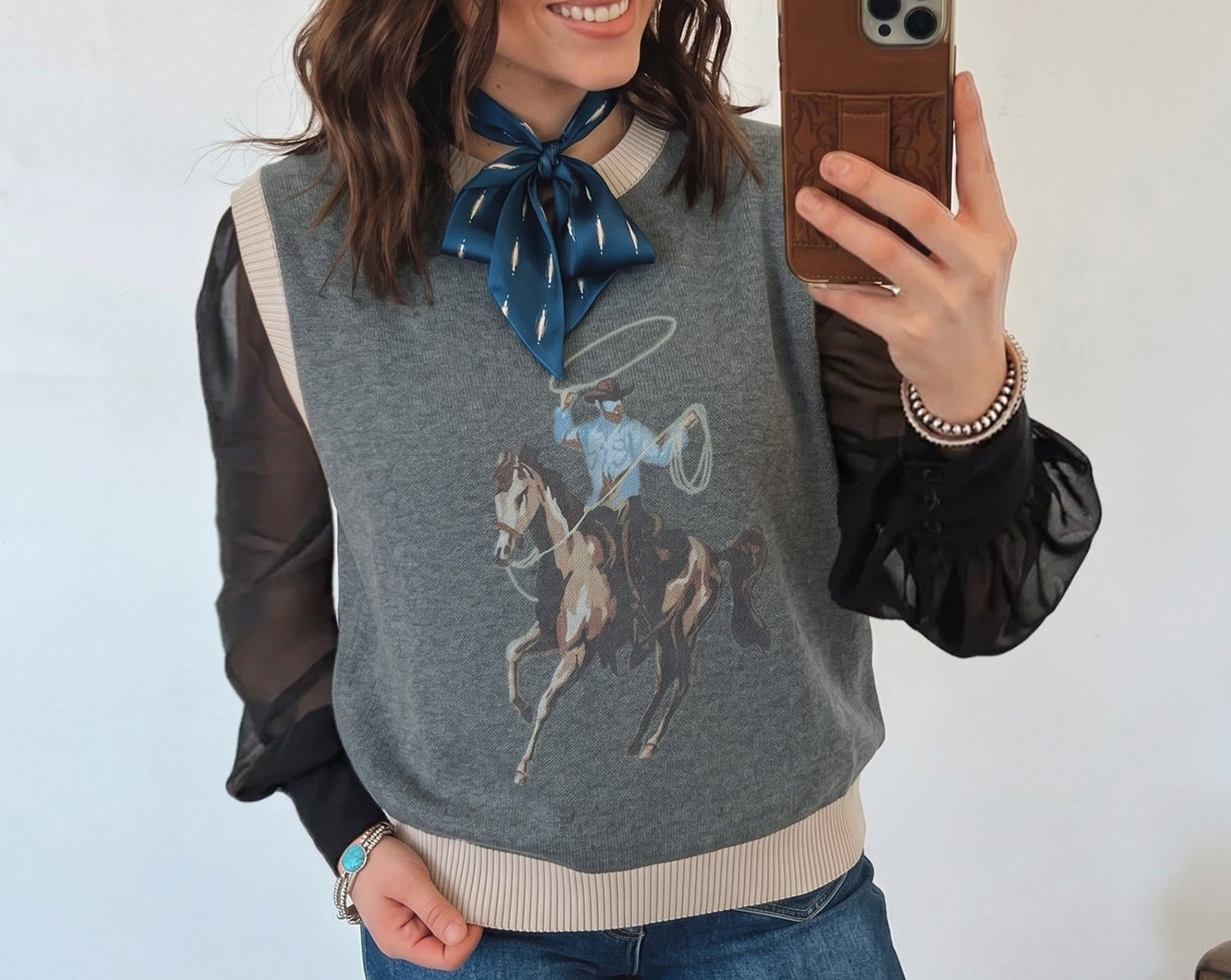Woman taking a mirror selfie wearing a gray sweater with a horse design, blue jeans, and a blue scarf.