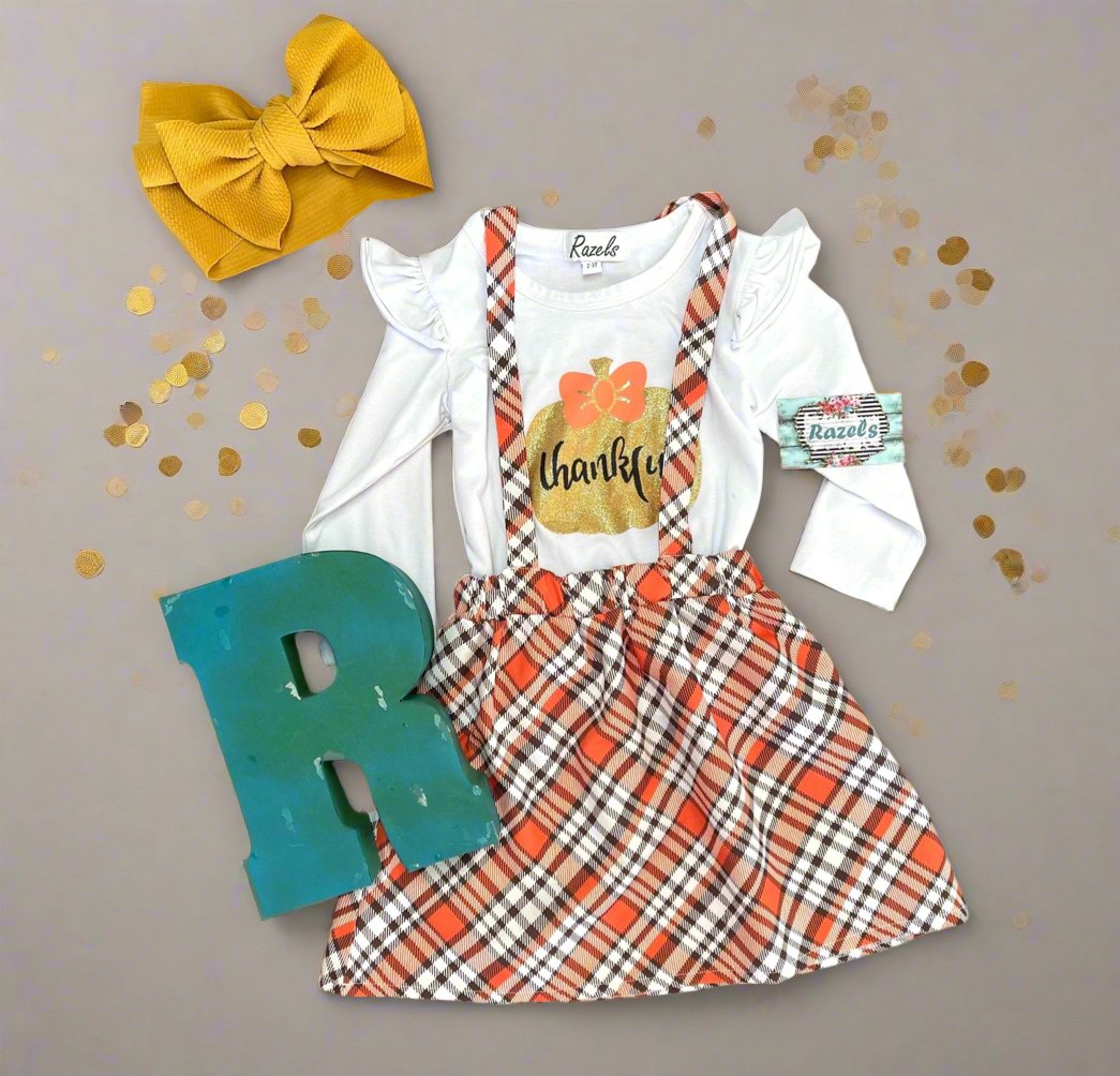 Girls' Fall Thankful Outfit. The Fall Plaid Suspender Skirt features an orange, brown, and white plaid pattern with suspenders. The shirt boasts ruffled shoulders, long sleeves, and a glittering gold pumpkin with the word 'thankful' in the center.