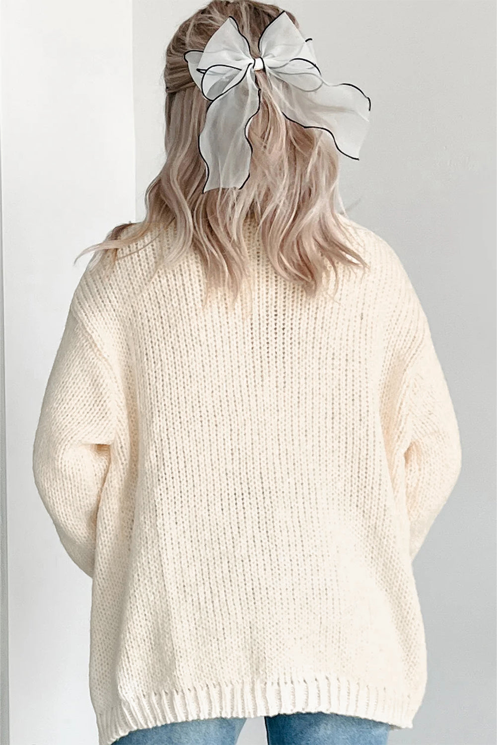 Person wearing a cream sweater with a white bow in their hair against a plain background