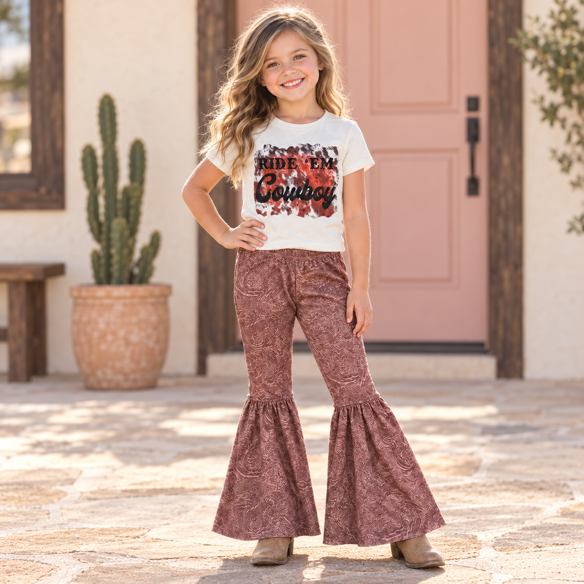 A young girl stands outdoors in front of a pink door and cactus planter, wearing a white graphic T-shirt that reads 'RIDE 'EM COWBOY' and dusty rose bell-bottom flare pants.
